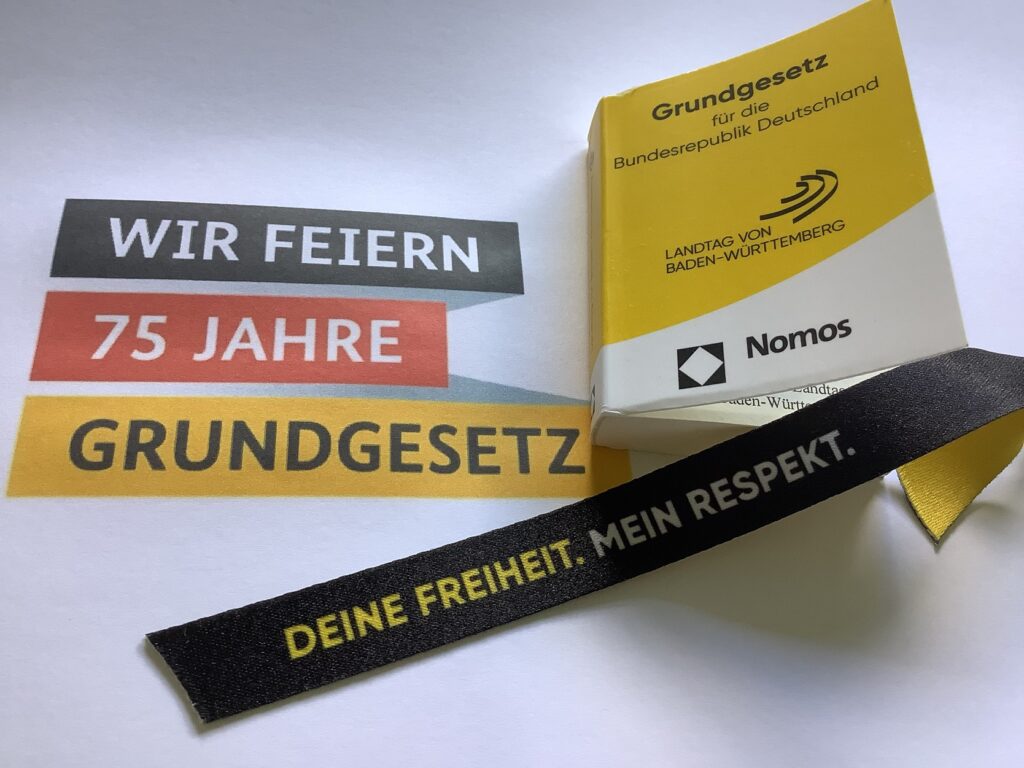 Ein deutsches Buch mit dem Titel „Grundgesetz für die Bundesrepublik Deutschland“ ist teilweise geöffnet. Daneben hängt ein Band mit der Aufschrift „Deine Freiheit. Mein Respekt.“ Auch auf dem Papier ist ein Motiv mit der Aufschrift „Wir feiern 75 Jahre Grundgesetz“ zu sehen.