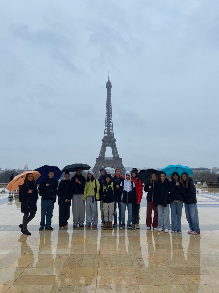 Eine Gruppe von Menschen mit Regenschirmen steht an einem bewölkten Tag auf einem nassen Platz vor dem Eiffelturm. Ihre Silhouetten spiegeln sich im Boden wider, und im Hintergrund ragt das berühmte Bauwerk empor.