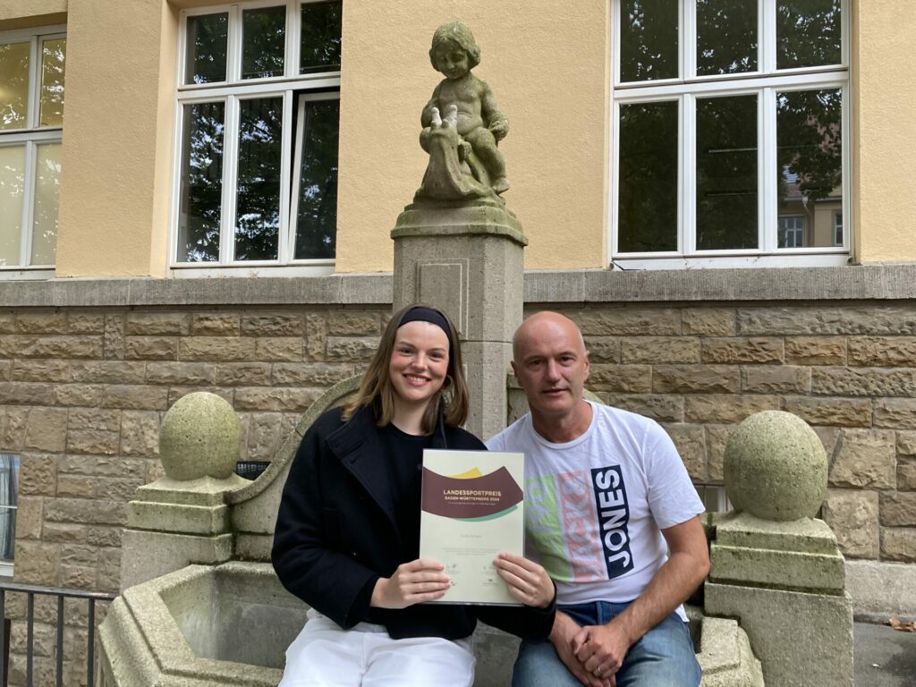Eine lächelnde Frau und ein Mann sitzen auf einer Steinbank vor einem Brunnen. Die Frau hält eine Urkunde in der Hand, und beide wirken zufrieden. Das Gebäude hinter ihnen hat große Fenster und Steinmauern.