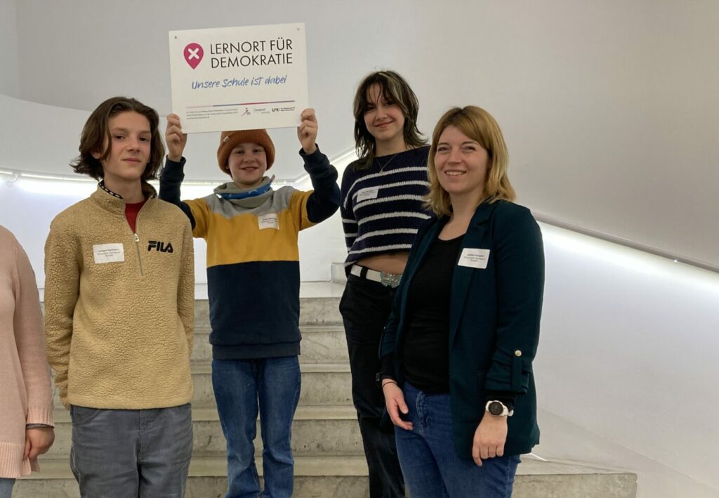 Vier Personen stehen auf einer Treppe, drei halten ein Schild mit der Aufschrift „Lernort für Demokratie“. Sie wirken fröhlich, alle tragen legere Kleidung. Die Szenerie wirkt wie ein modern gestalteter Innenraum.