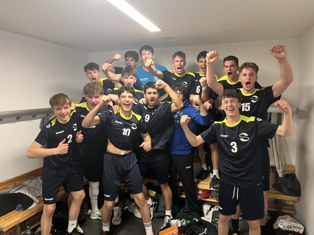 Eine Gruppe von Teenagern in passenden dunklen Sportuniformen feiert aufgeregt in einer Umkleidekabine, hebt die Fäuste und lächelt. Einige stehen auf Bänken, und um sie herum sind Sportgeräte zu sehen.