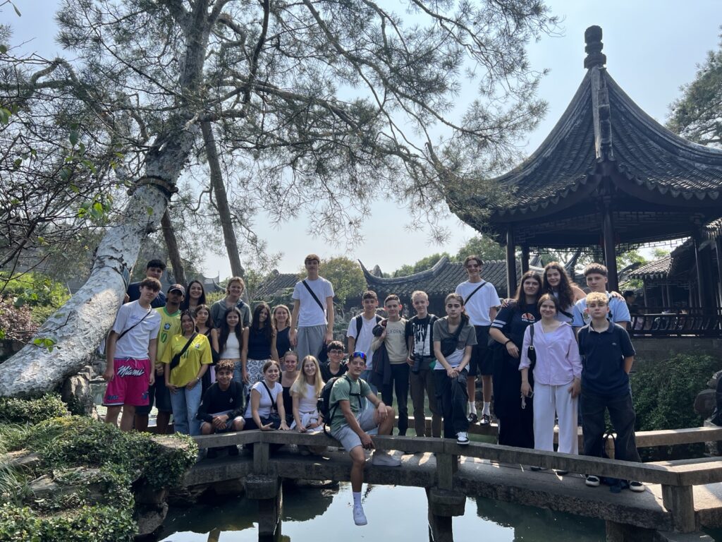 Eine Gruppe von Jugendlichen und Erwachsenen posiert für ein Foto auf einer Holzbrücke in einem traditionellen chinesischen Garten mit einem Teich, üppigem Grün und einem Pavillon im Hintergrund unter einem sonnigen Himmel.