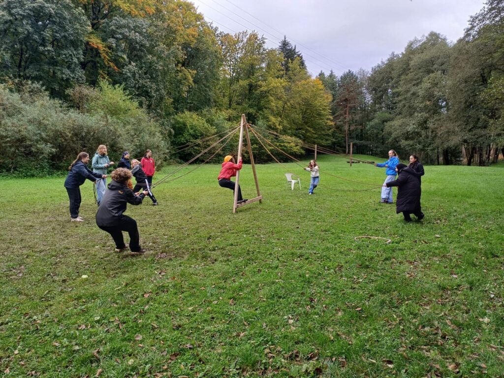 Eine Gruppe von Menschen in einem Park hält Seile an einer Holzkonstruktion fest und balanciert jemanden in der Mitte. Der Boden ist grasbewachsen, umgeben von Bäumen, und der Himmel ist bedeckt. Es scheint sich um eine Teambuilding-Aktivität zu handeln.