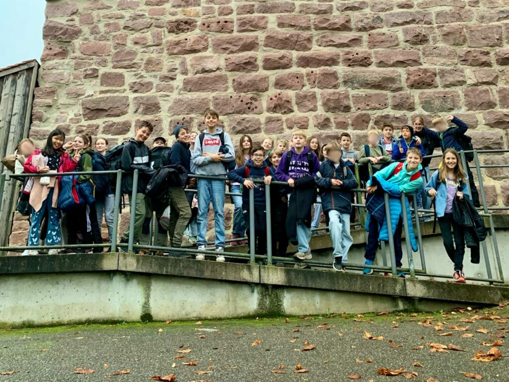 Eine Gruppe Kinder und einige Erwachsene stehen auf einer Rampe vor einem Steingebäude. Sie lächeln und scheinen einen Ausflug zu machen. Der Boden ist nass und mit Blättern bedeckt. Einige tragen Taschen und Jacken.