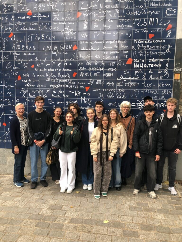 Eine Gruppe von elf Personen posiert lächelnd vor der "I Love You"-Mauer in Paris, die mit den Worten "Ich liebe dich" in vielen verschiedenen Sprachen bedeckt ist.
