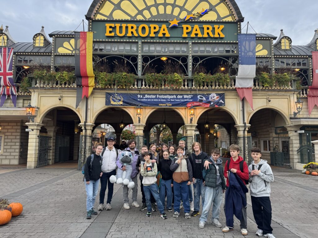 Eine Gruppe von Menschen, darunter mehrere Kinder, steht lächelnd vor dem Eingang des Europa-Parks. Sie posieren an einem bewölkten Tag für ein Foto. Einige halten Stofftiere in den Händen, und am Gebäude sind Fahnen zu sehen.