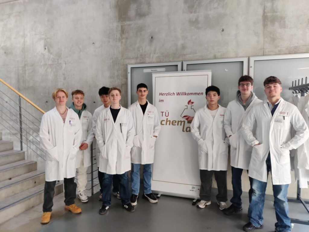 Eine Gruppe von acht Personen in weißen Laborkitteln steht vor einem Schild mit der Aufschrift „Herzlich Willkommen im TU-Chem-Labor“ in einem modernen Gebäude mit einer Betonwand und einer Treppe auf der linken Seite.