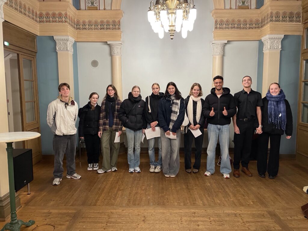 Eine Gruppe von zehn Personen steht in einem Raum mit kunstvollen architektonischen Details. Sie lächeln in die Kamera, sind leger gekleidet und halten Papiere in der Hand. Der Raum ist mit Holzböden und einem dekorativen Kronleuchter ausgestattet.