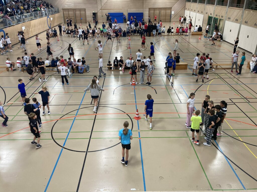 Eine große Turnhalle, in der sich Menschen unterschiedlichen Alters tummeln. Kindergruppen spielen und beschäftigen sich auf dem Platz. Zuschauer sitzen und stehen an den Seiten. Die Atmosphäre ist lebhaft und energiegeladen.