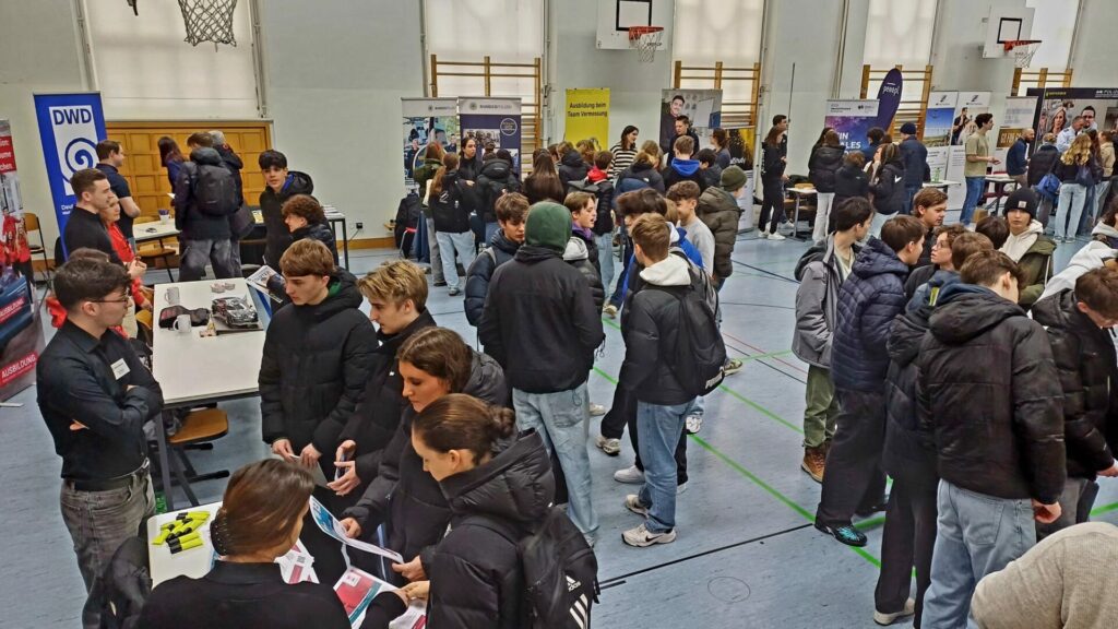In einer überfüllten Turnhalle findet eine Karrieremesse mit verschiedenen Ständen und Bannern statt. Menschen, meist junge Erwachsene in Winterkleidung, unterhalten sich mit Vertretern, halten Broschüren in den Händen und diskutieren über Möglichkeiten. Im Hintergrund sind Basketballkörbe zu sehen.