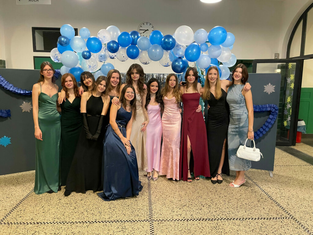 Eine Gruppe von elf jungen Frauen in Abendkleidern posiert vor einem festlichen Ballonbogen. Die Kleider variieren in den Farben Schwarz, Rot und Pink. Im Hintergrund sind blaue und weiße Ballons und dekorative Sterne zu sehen.