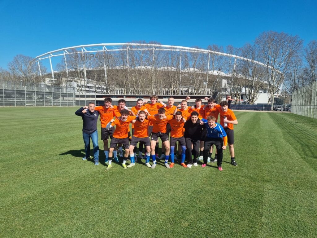 Eine Gruppe Fußballspieler in orangefarbenen Trikots und schwarzen Shorts posiert fröhlich auf einem Rasenplatz. Sie stehen Arm in Arm vor einem Stadion unter einem strahlend blauen Himmel.