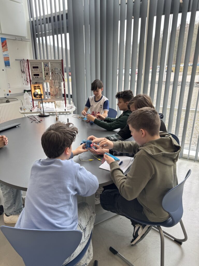 Eine Gruppe Jungen sitzt um einen Tisch und arbeitet an einem praktischen Wissenschaftsprojekt. Sie bauen Schaltkreise mit Drähten und Geräten zusammen. Der Raum hat große Jalousien an den Fenstern und lehrreiche Poster an den Wänden.