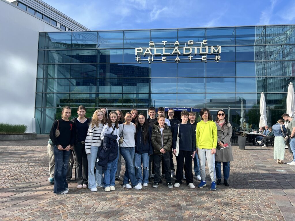 Eine Gruppe Teenager und zwei Erwachsene stehen lächelnd vor dem Stage Palladium Theater, mit einer modernen Glasfassade und blauem Himmel im Hintergrund.