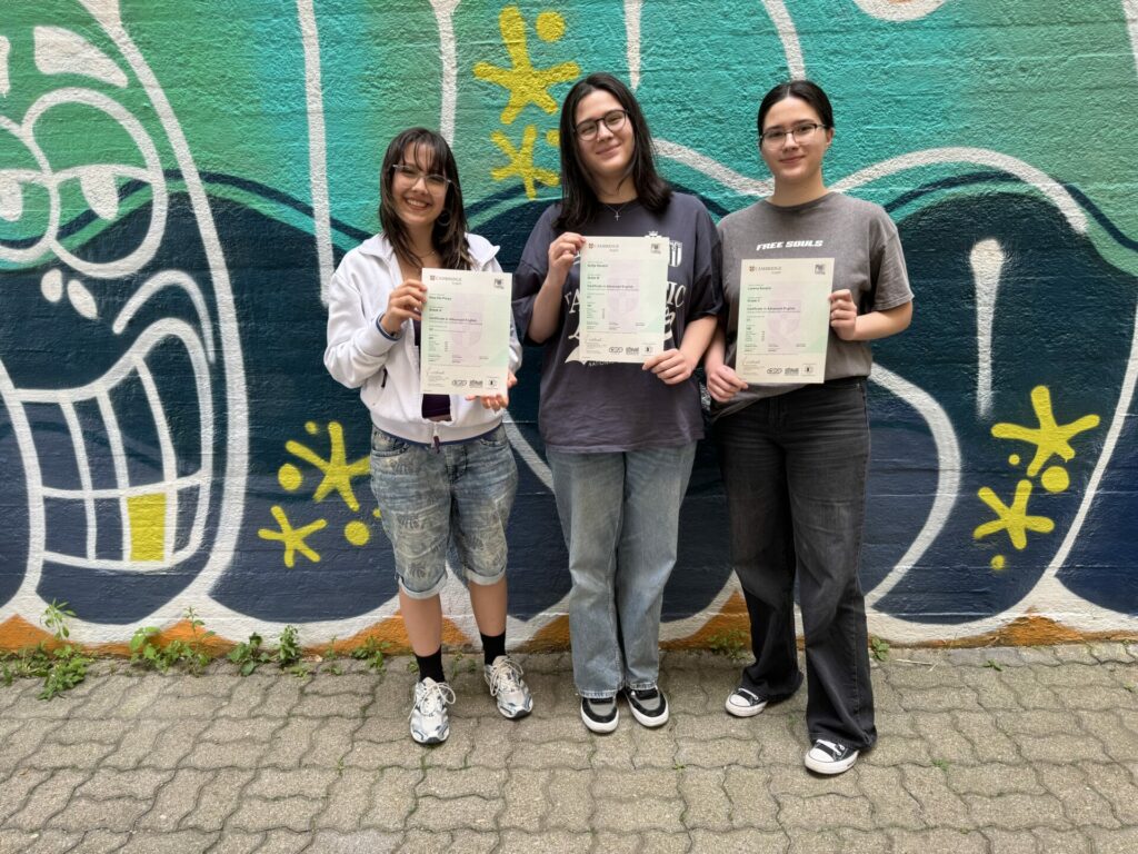 Drei junge Menschen stehen lächelnd vor einer bunten Graffiti-Wand und halten Zertifikate in den Händen. Sie tragen Freizeitkleidung und stehen auf einem gepflasterten Weg, der von Grünpflanzen gesäumt ist.