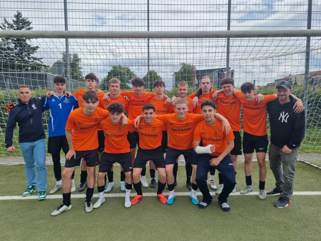 Eine Gruppe junger Männer in orangefarbenen Fußballtrikots posiert gemeinsam vor einem Tor auf einem Außenfeld. An beiden Enden stehen zwei Erwachsene. Das Team lächelt für das Foto. Im Hintergrund sind Grünflächen und Gebäude zu sehen.