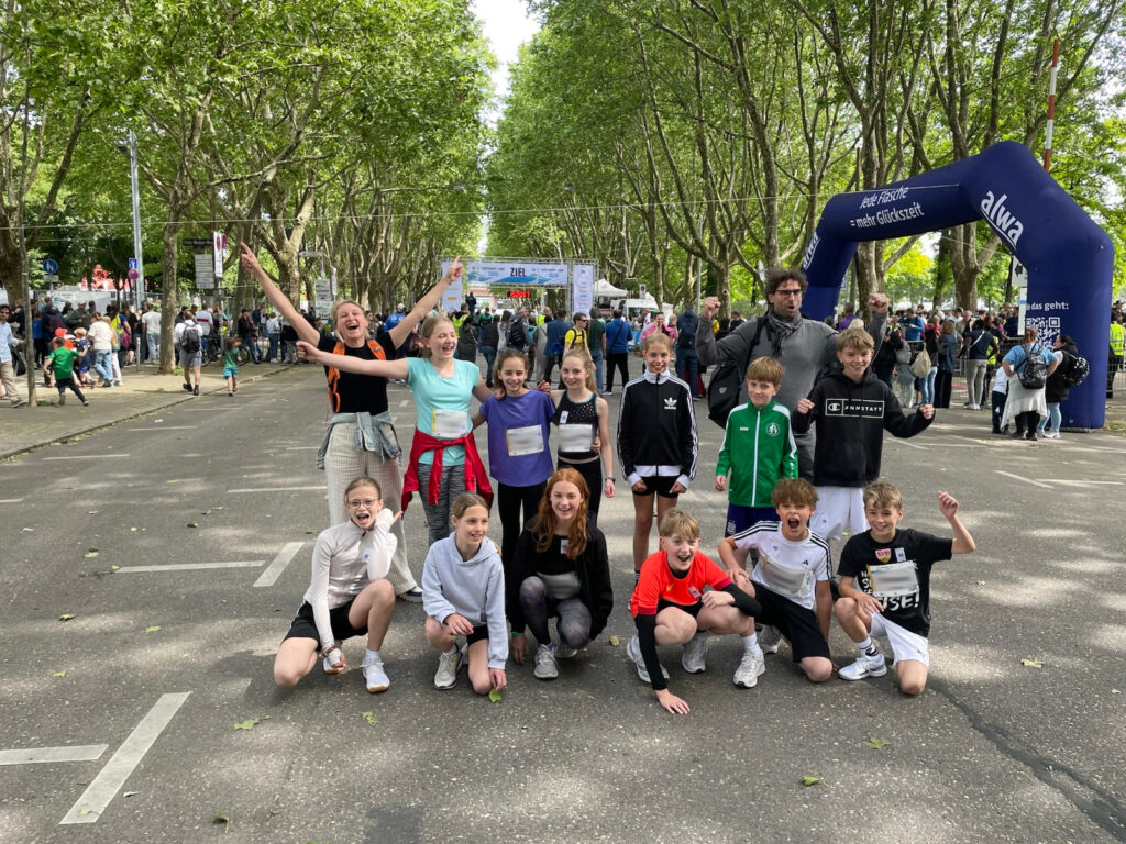 Eine Gruppe lächelnder Kinder und ein Erwachsener posieren gemeinsam auf einer Straße bei einer Veranstaltung im Freien, mit vielen Menschen und Bäumen im Hintergrund, in der Nähe eines blauen aufblasbaren Bogens.