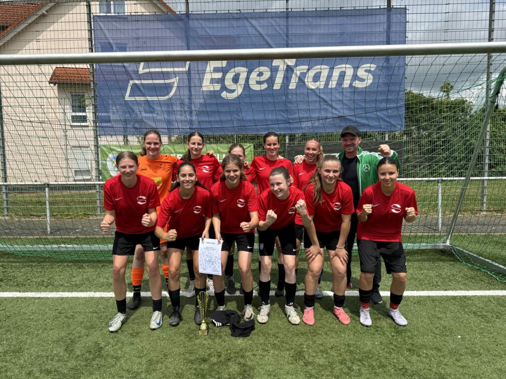 Eine Mädchenfußballmannschaft in roten Trikots posiert lächelnd vor einem Tor und feiert mit Pokal und Urkunde einen Sieg. Ein Trainer steht mit ihnen unter einem EgeTrans-Banner.