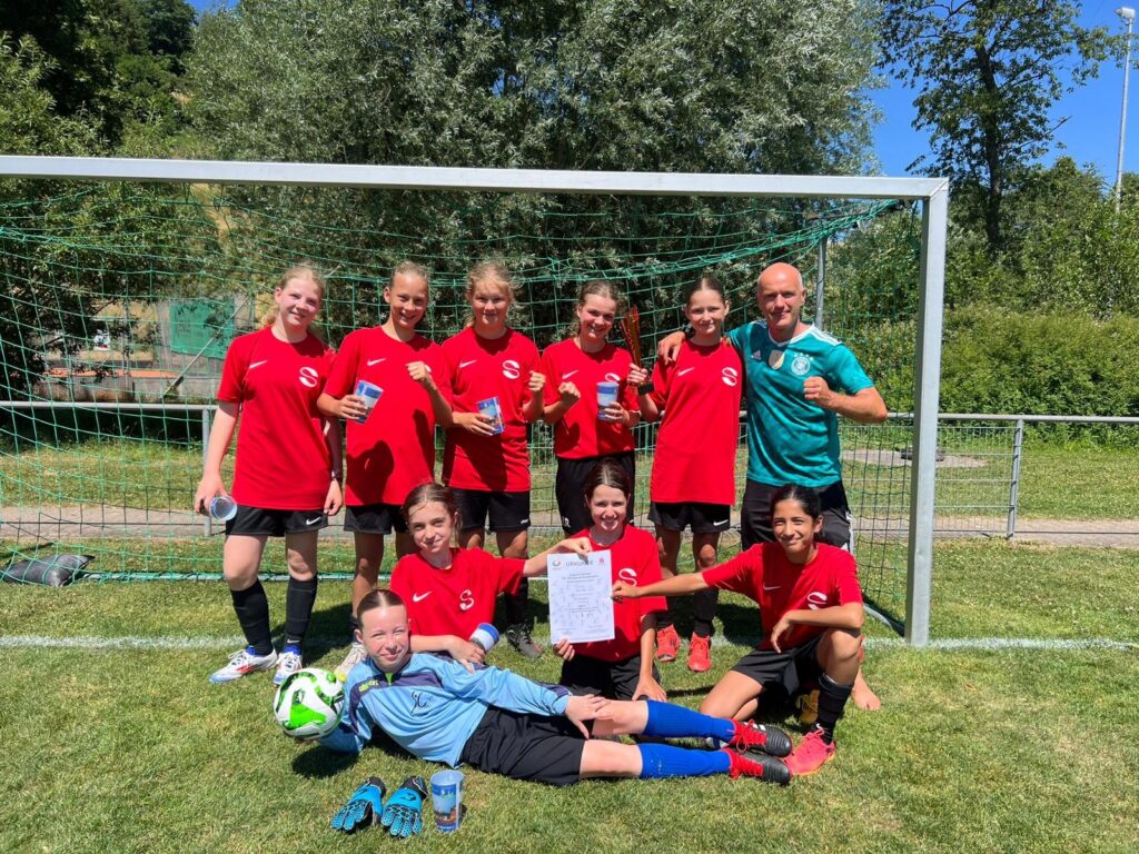 Eine Mädchenfußballmannschaft in roten Trikots posiert fröhlich mit ihrem Trainer vor einem Tor. Ein Mädchen hält eine Urkunde in der Hand, ein anderes einen Fußball, und ein Spieler in Torwartkleidung liegt vor der Gruppe.