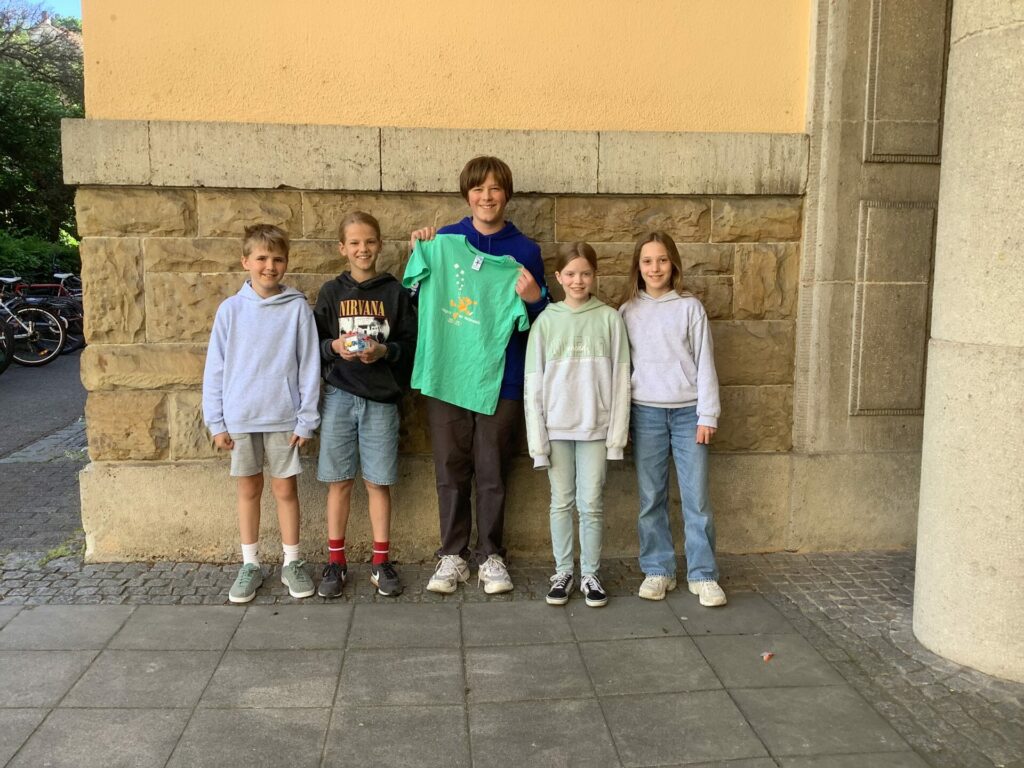 Fünf Kinder stehen nebeneinander vor einer Steinmauer. Das mittlere Kind hält ein grünes T-Shirt hoch. Alle lächeln und sind leger gekleidet. Zwei tragen Shorts, drei Hosen.