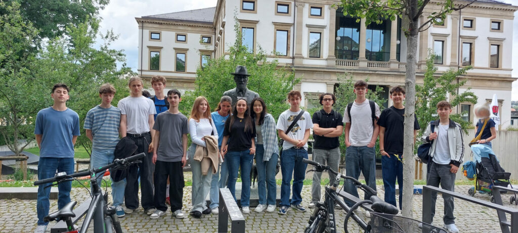 Eine Gruppe von dreizehn jungen Leuten steht draußen vor einem großen Gebäude mit hohen Fenstern, hinter ihnen eine Statue und im Vordergrund mehrere geparkte Fahrräder.