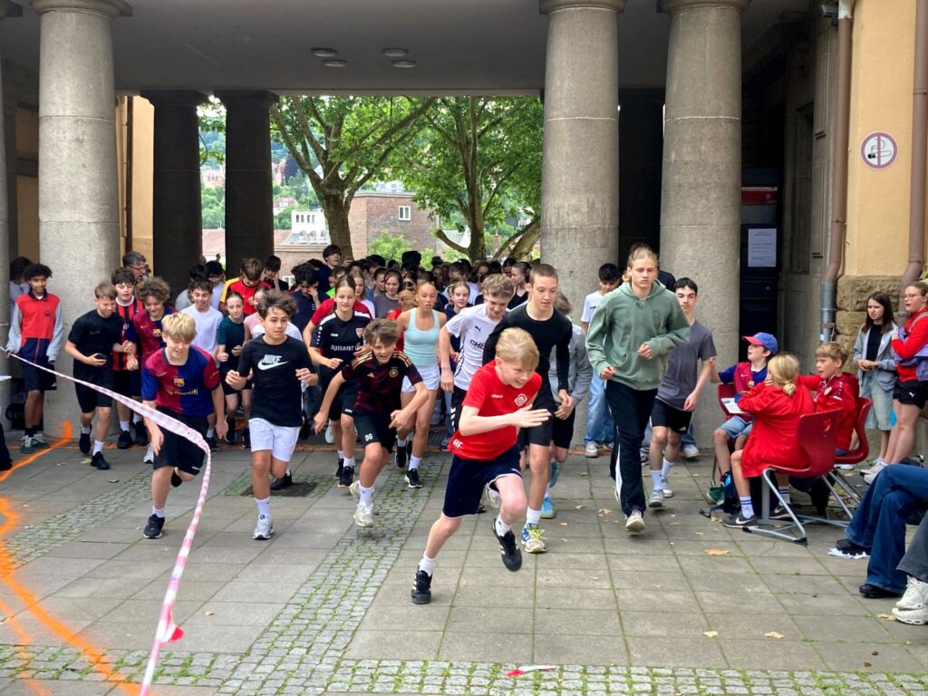 Eine große Gruppe von Kindern und Jugendlichen, einige in Sportkleidung, rennt von einem Hof unter einem Torbogen los; Erwachsene und andere schauen von der Seite zu und jubeln. Im Hintergrund sind Bäume und Gebäude zu sehen.