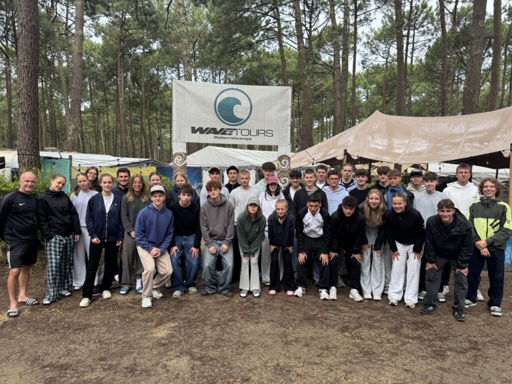 Eine große Gruppe von Menschen, hauptsächlich Teenager, posiert und lächelt vor einem Wavetours-Banner in einem Outdoor-Camp, umgeben von hohen Kiefern und Zelten.