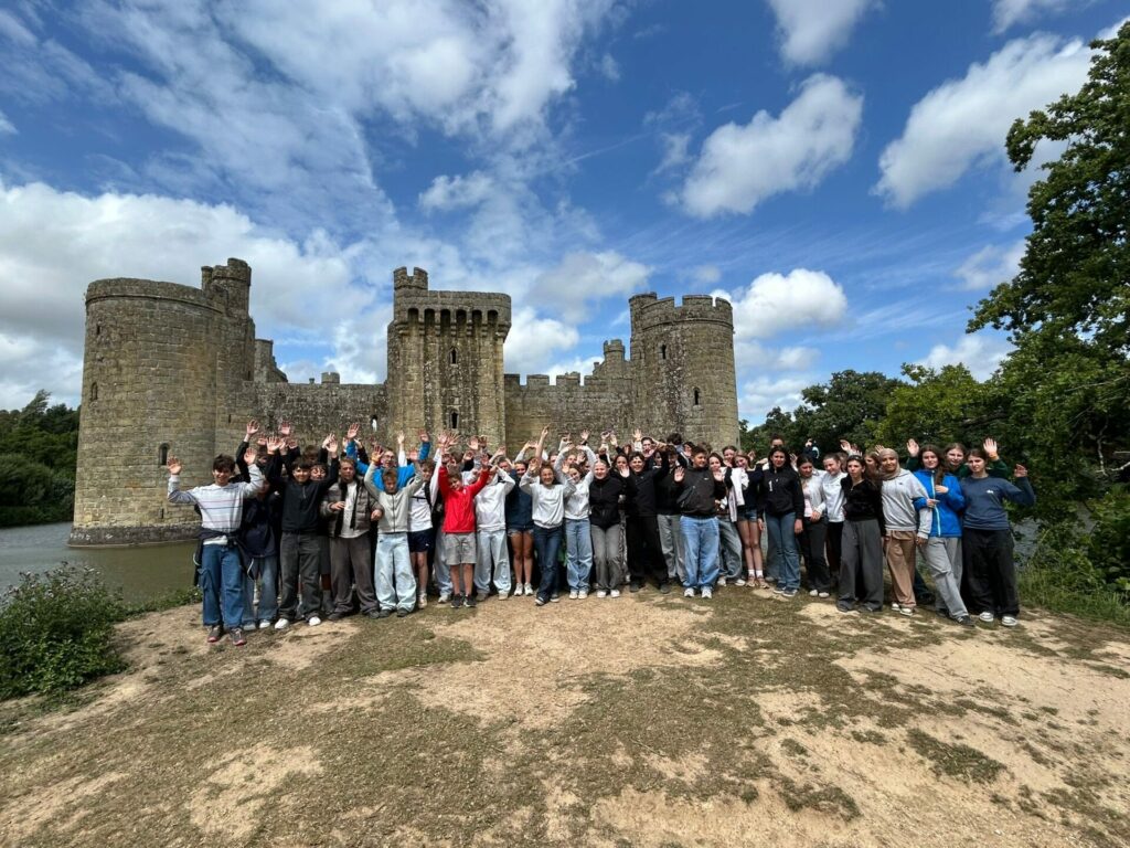 Eine große Gruppe von Menschen steht lächelnd mit erhobenen Armen vor einer historischen Steinburg, umgeben von Grün, unter einem teilweise bewölkten blauen Himmel.