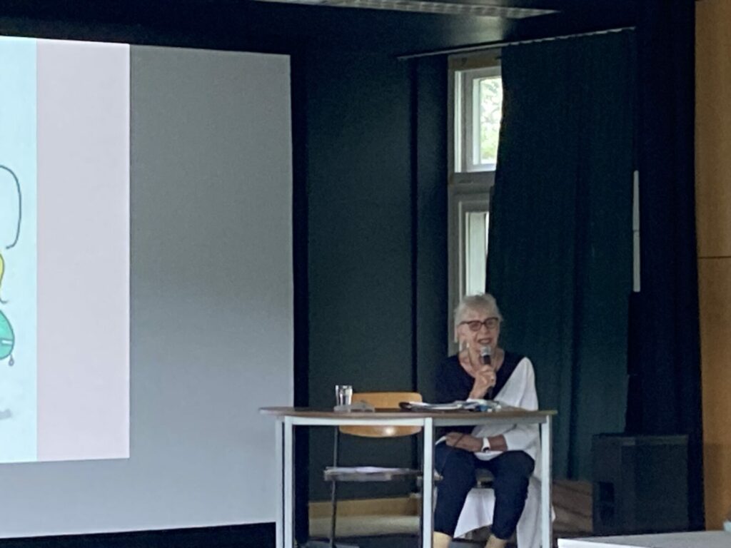 Eine ältere Frau sitzt an einem Schreibtisch mit einem Mikrofon und spricht in einem schwach beleuchteten Raum vor einer Projektionsfläche. Ein Glas Wasser und Papiere stehen auf dem Schreibtisch. Im Hintergrund befinden sich Vorhänge und ein Fenster.