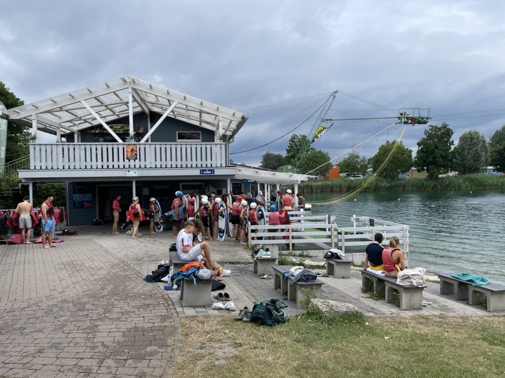 An einem bewölkten Tag versammeln sich Menschen in der Nähe eines Gebäudes am See, einige in Badekleidung, andere bereiten sich auf Wasseraktivitäten vor. Wakeboarding-Ausrüstung ist über dem Wasser zu sehen, und Handtücher und Taschen liegen auf Bänken verstreut.
