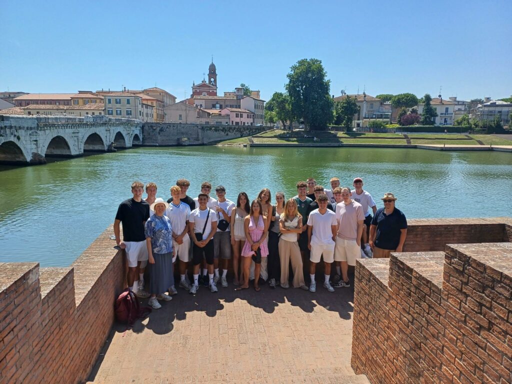 Eine Gruppe von Menschen posiert für ein Foto auf einer Ziegelsteinbrücke an einem Fluss, mit historischen Gebäuden und einer Steinbrücke im Hintergrund unter einem sonnigen, klaren blauen Himmel.