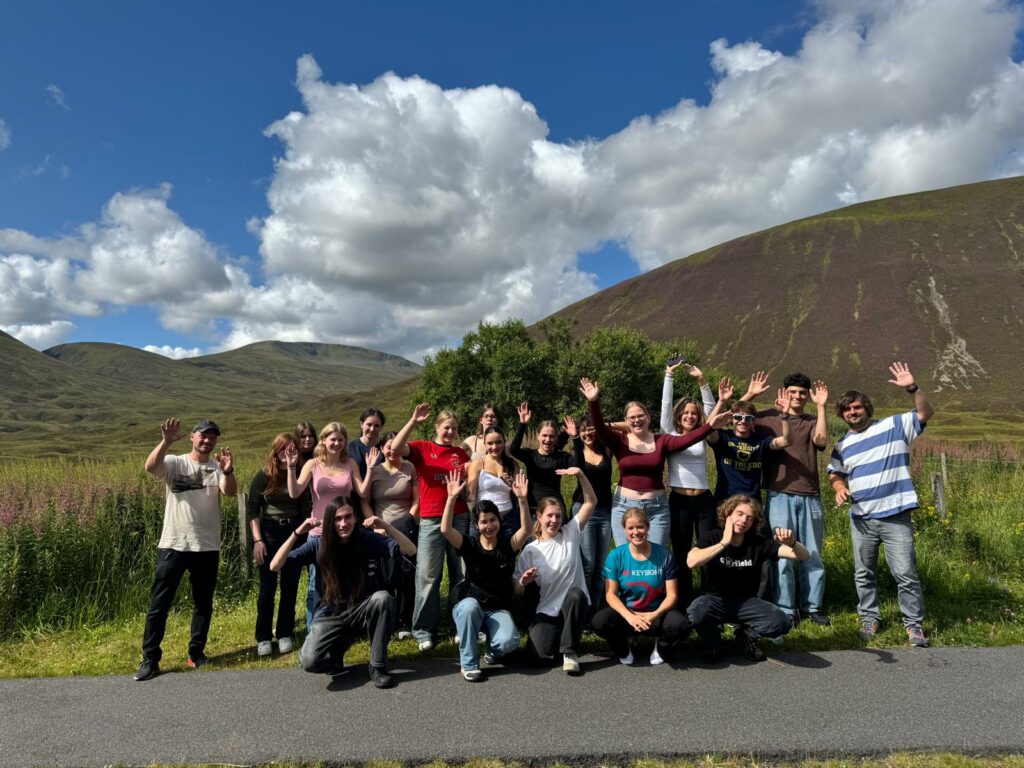 Eine Gruppe von Menschen posiert an einem sonnigen Tag im Freien, lächelnd und winkend mit Hügeln, Gras und einem blauen Himmel mit Wolken im Hintergrund.
