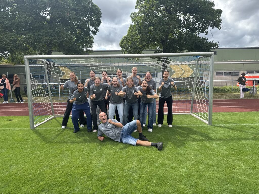 Eine Gruppe von Personen in passenden grauen Hemden posiert aufgeregt vor einem Fußballtor auf einer Wiese, wobei eine Person davor liegt. Im Hintergrund sind bei bewölktem Himmel Bäume und eine Rennstrecke zu sehen.