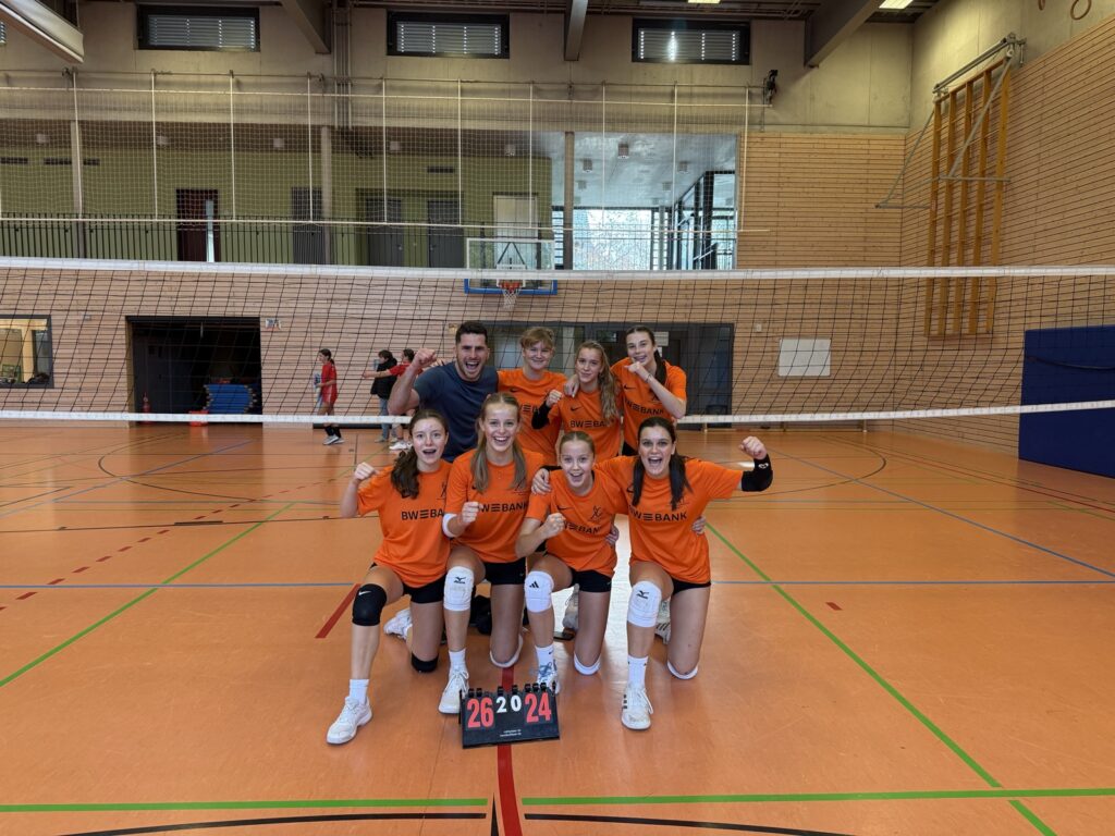 Acht Volleyballspieler in orangefarbenen Trikots feiern auf einem Hallenplatz, kniend und stehend hinter einer Anzeigetafel, die ein Ergebnis von 26:24 anzeigt. Sie lächeln und machen triumphierende Gesten.