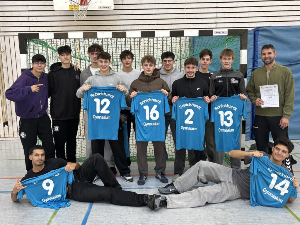 Eine Gruppe von Teenagern und ein Erwachsener stehen und sitzen in einer Turnhalle und posieren mit blauen Sporttrikots mit den Nummern 9, 12, 14, 16, 2 und 13. Der Erwachsene hält eine Urkunde in der Hand. Im Hintergrund sind ein Netz und eine Holzwand zu sehen.