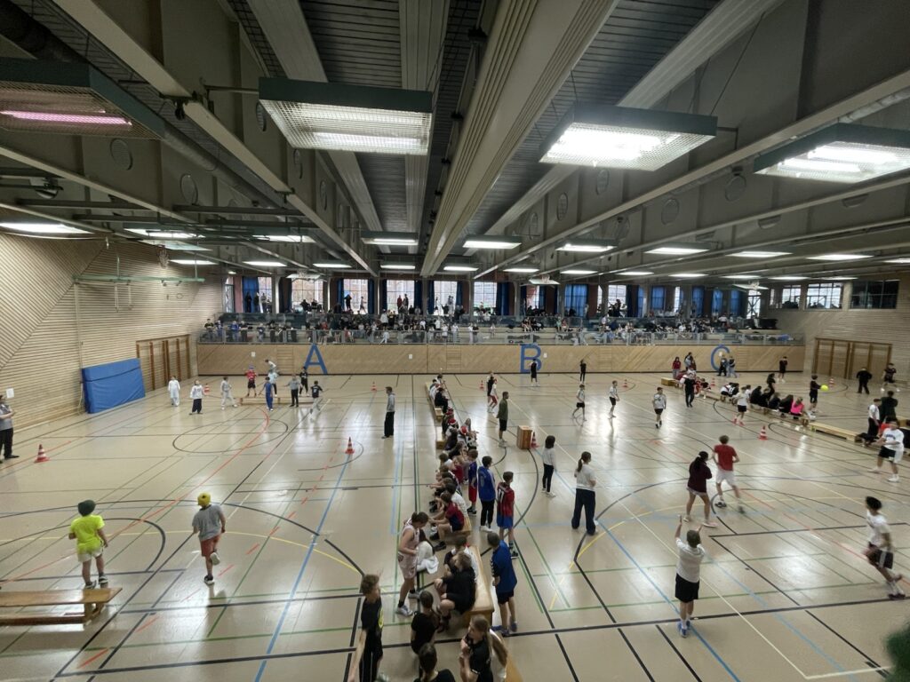 Eine große, überdachte Sporthalle, in der Menschen an verschiedenen sportlichen Aktivitäten teilnehmen. Die Zuschauer sitzen auf Tribünen an der Rückwand. Der Boden der Halle ist mit verschiedenfarbigen Linien markiert, und es sind mehrere Kegel und Geräte zu sehen.