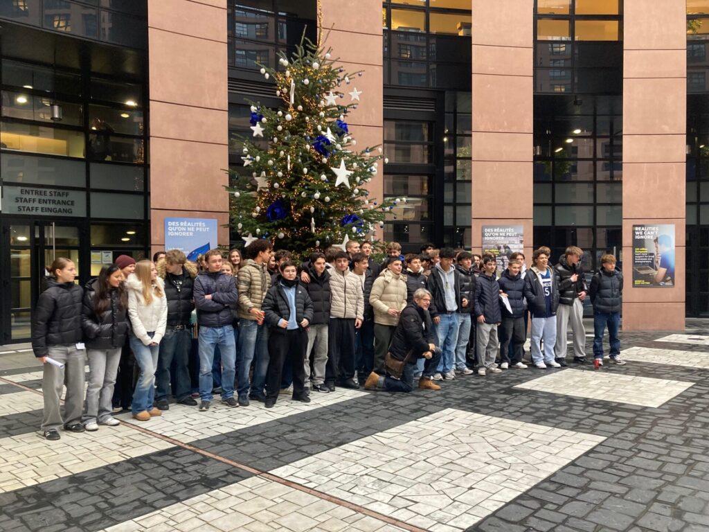 Eine große Gruppe von Menschen steht vor einem geschmückten Weihnachtsbaum im Freien, umgeben von hohen Gebäuden mit Glasfenstern und Plakaten an den Wänden. Viele tragen Wintermäntel.