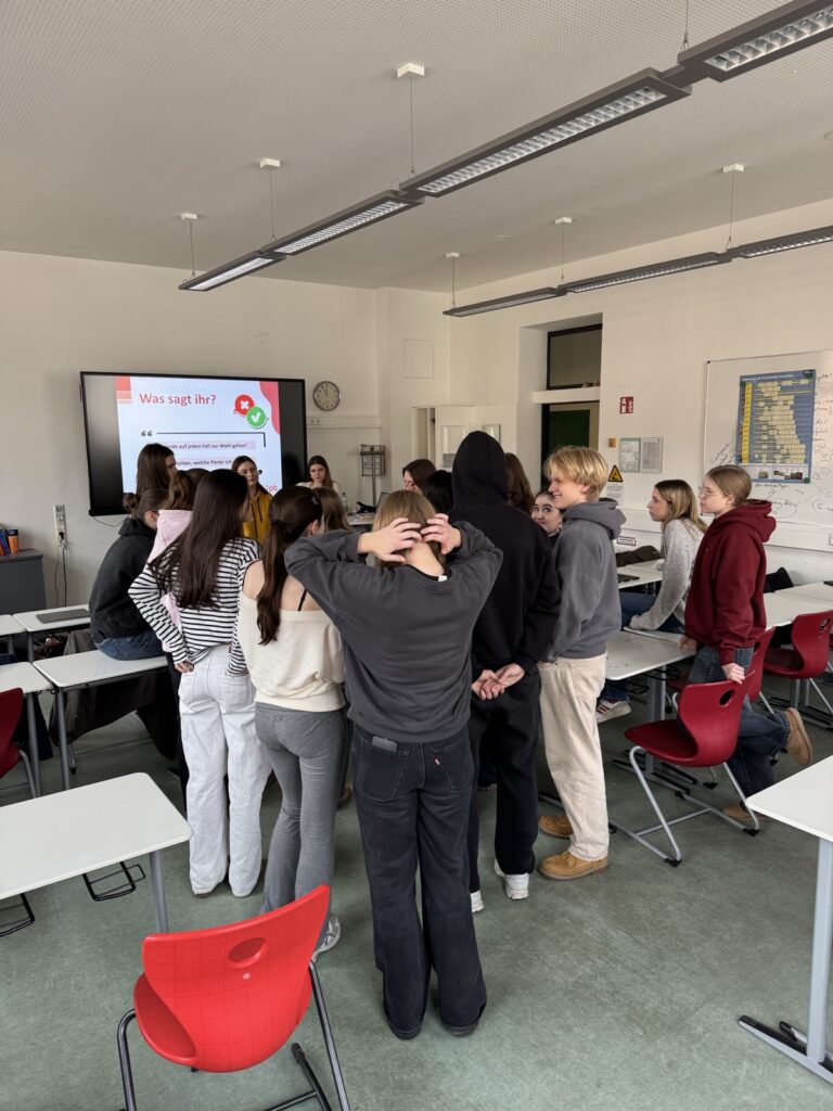 Eine Gruppe von Schülern steht in einem Klassenzimmer im Kreis und blickt auf einen Präsentationsbildschirm an der Stirnseite. Auf dem Bildschirm wird ein Text in deutscher Sprache angezeigt. Tische und rote Stühle sind im Raum verteilt.