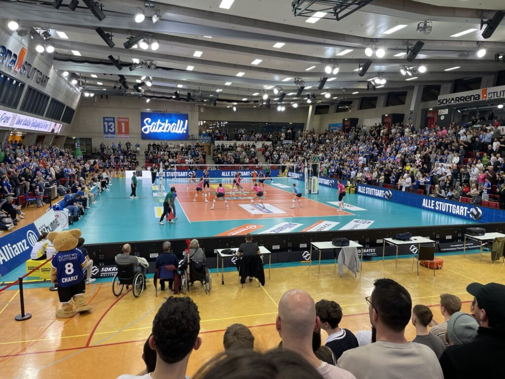 Eine große Volleyballhalle, gefüllt mit Zuschauern, die ein Spiel verfolgen. Die Spieler sind auf dem Spielfeld, und ein Maskottchen in einem Trikot steht in der Nähe von Menschen in Rollstühlen am Eingang. Helle Lichter erhellen die Szene.