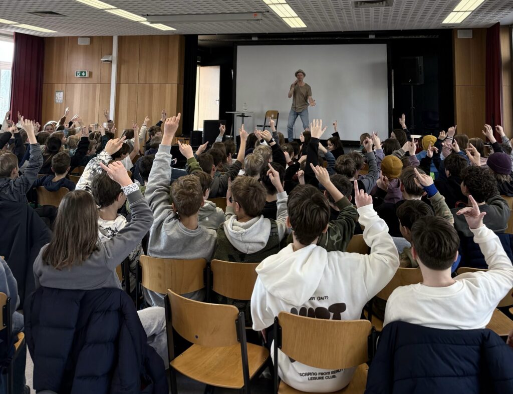 Eine große Gruppe von Schülerinnen und Schülern sitzt vor einer Bühne und hebt die Hände, während ein Moderator mit Hut über ein Mikrofon vor einer Projektionsfläche in einer Schulaula zu ihnen spricht.