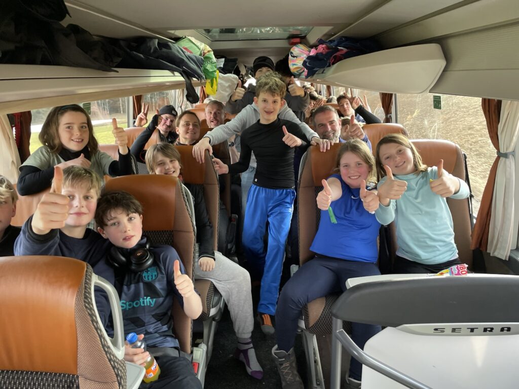 Eine Gruppe lächelnder Kinder und Jugendlicher sitzt und steht in einem Bus und zeigt mit dem Daumen nach oben in die Kamera. Die Atmosphäre ist fröhlich und energiegeladen, mehrere Kinder tragen legere Kleidung und einige haben Snacks oder Getränke in der Hand.