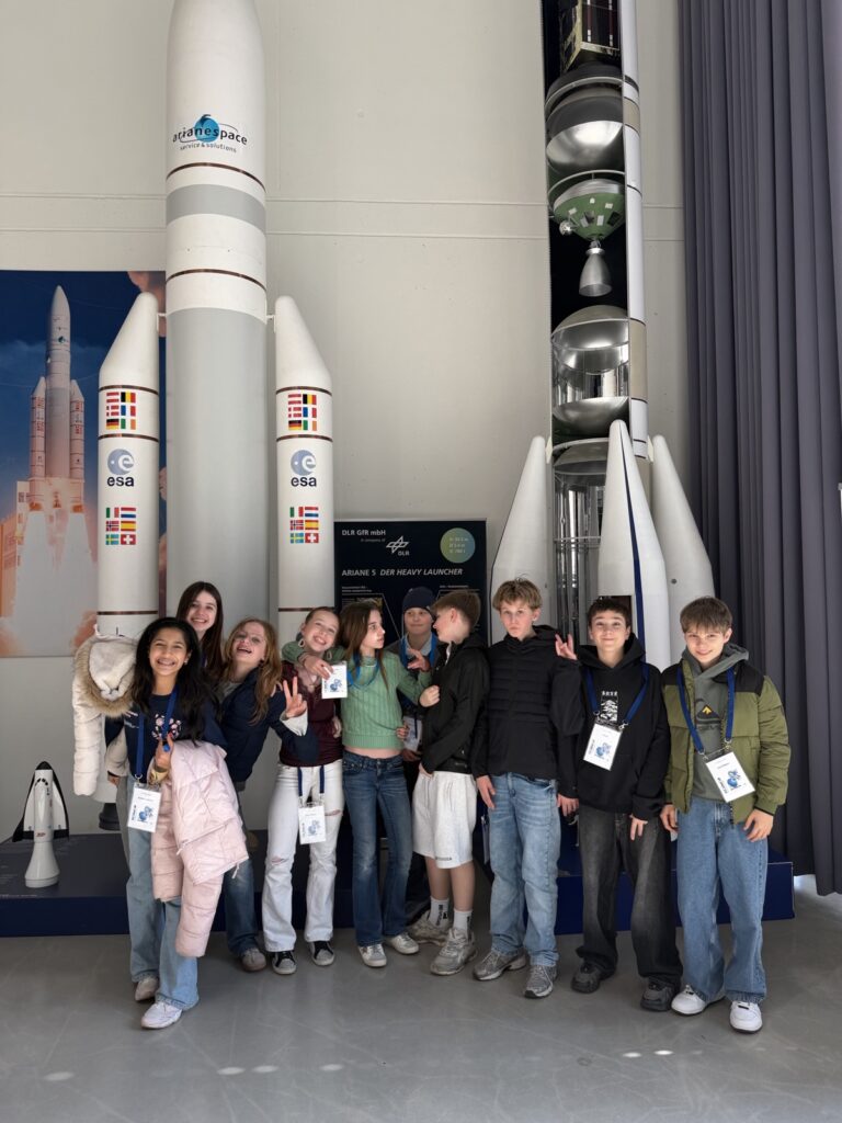 Eine Gruppe von zehn lächelnden Teenagern posiert vor großen ESA-Modellraketen und Schautafeln in einer Raumfahrtausstellung und trägt Besucherausweise um den Hals.