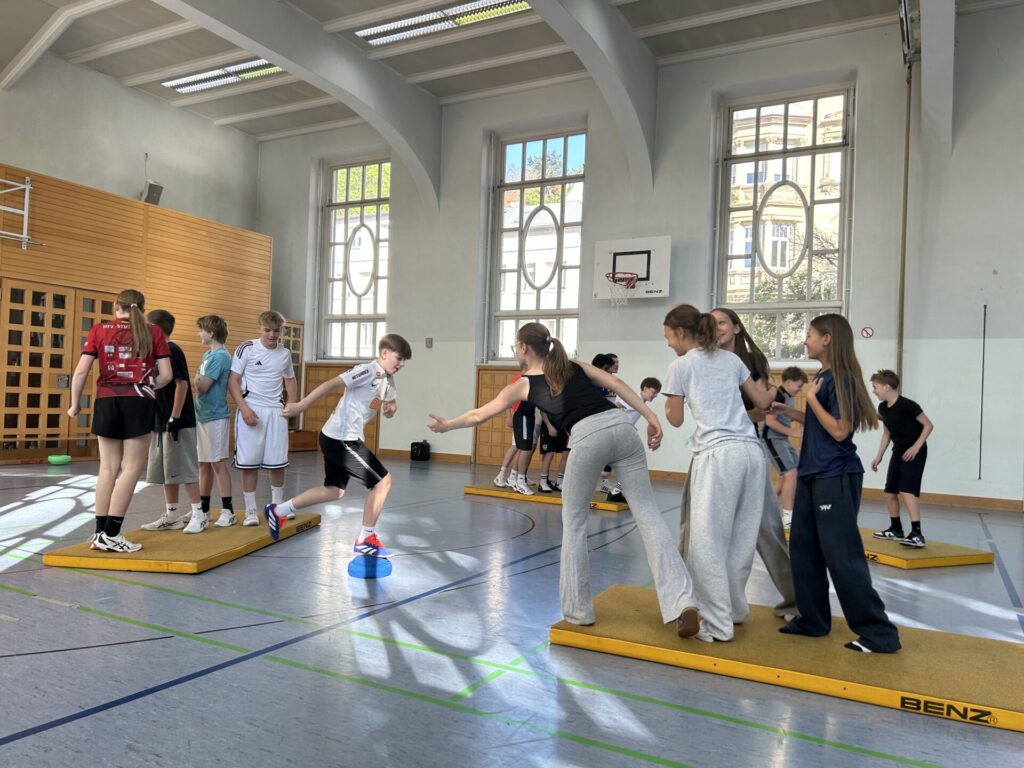 Eine Gruppe von Kindern in Sportkleidung nimmt an einer Teamaktivität auf Matten in einer Turnhalle teil, wobei sie sich die Hände reichen, um sich gegenseitig zu markieren, wobei große Fenster natürliches Licht hereinlassen.