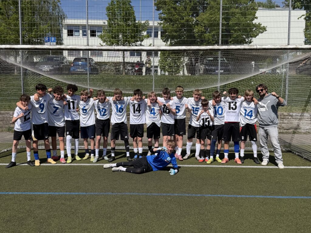 Eine Jugendfußballmannschaft in weißen Trikots posiert vor einem Tor auf einem Außenplatz. Ein Spieler in einem blauen Torwarttrikot liegt davor auf dem Boden, während ein Trainer im Trainingsanzug rechts daneben steht und lächelt. Es ist ein sonniger Tag.