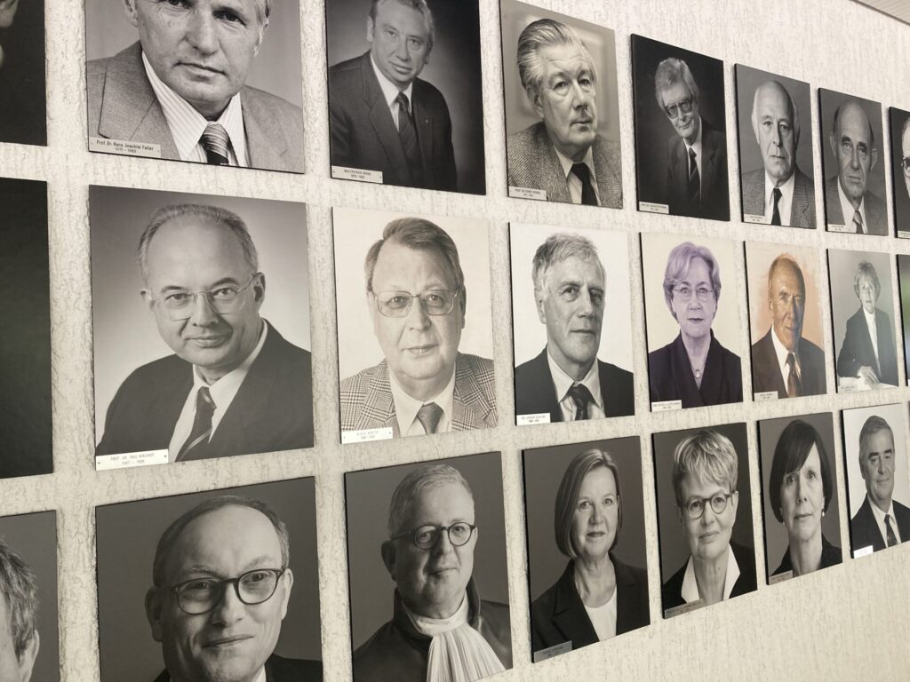 Eine Wand mit Schwarz-Weiß-Porträtfotos von verschiedenen Männern und Frauen, die in Reihen angeordnet sind. Die Porträts wirken förmlich und zeigen Personen in Berufskleidung, möglicherweise Richter oder Beamte.