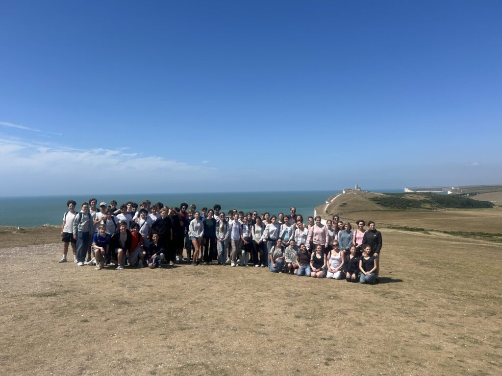 Eine große Gruppe von Menschen posiert gemeinsam auf einem grasbewachsenen Hügel vor dem Hintergrund des Meeres und des blauen Himmels, in der Nähe weißer Klippen und eines in der Ferne liegenden Leuchtturms.