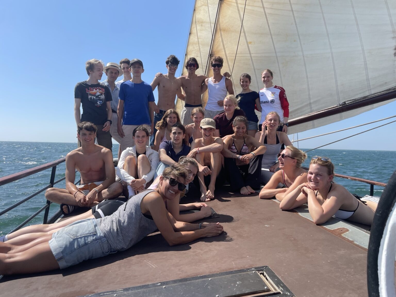 Eine Gruppe junger Leute, von denen einige Badeanzüge tragen, posiert und lächelt auf dem Deck eines Segelboots unter strahlendem Himmel, mit dem Meer im Hintergrund.