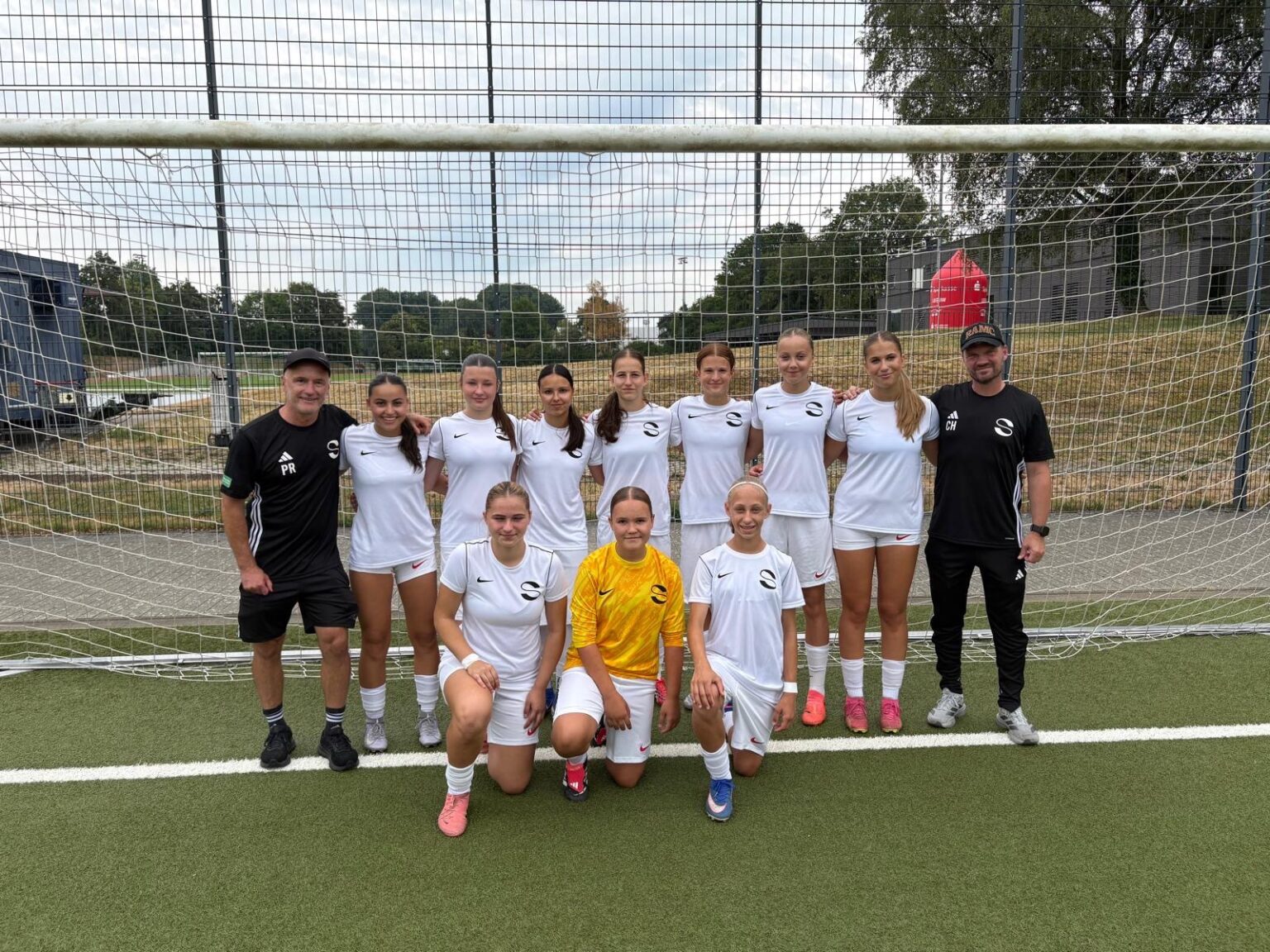 Eine Frauenfußballmannschaft posiert vor einem Tor auf einem Spielfeld. Die meisten Spielerinnen tragen weiße Trikots, eine trägt ein gelbes. Zwei schwarz gekleidete Trainer stehen zu beiden Seiten. Im Hintergrund sind Bäume und ein Zaun zu sehen.