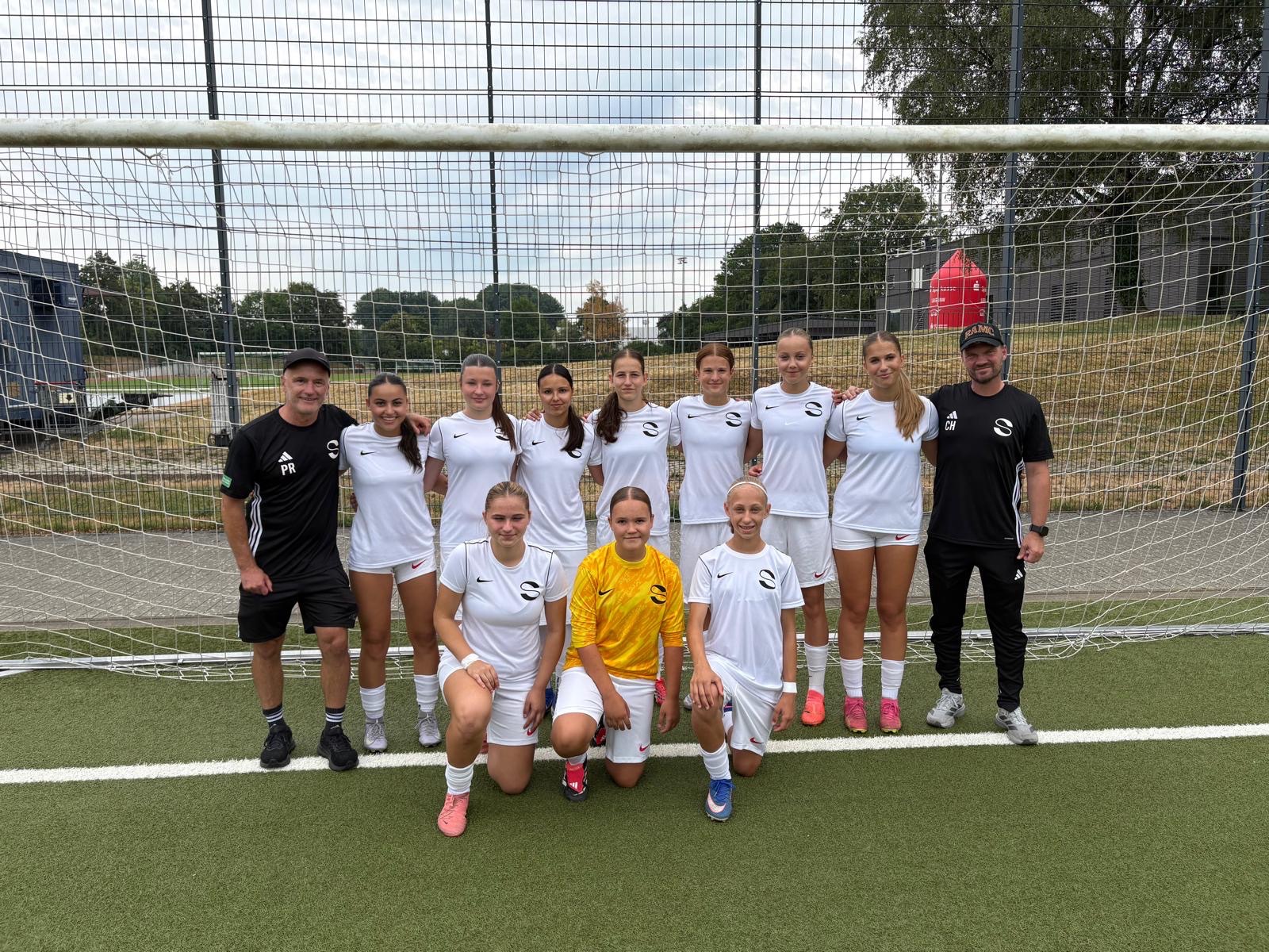 Eine Frauenfußballmannschaft posiert vor einem Tor auf einem Spielfeld. Die meisten Spielerinnen tragen weiße Trikots, eine trägt ein gelbes. Zwei schwarz gekleidete Trainer stehen zu beiden Seiten. Im Hintergrund sind Bäume und ein Zaun zu sehen.