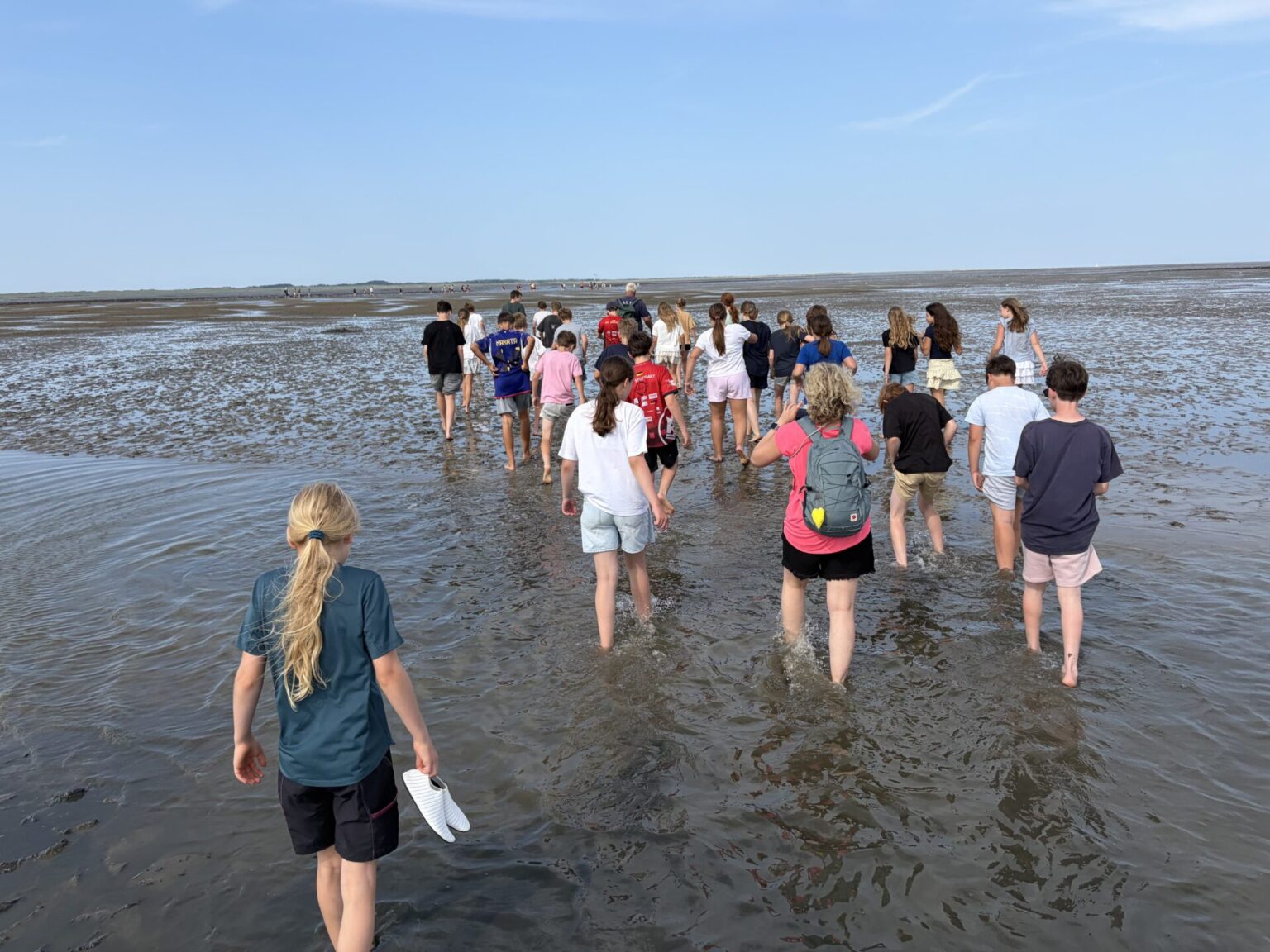 Eine Gruppe von Menschen, hauptsächlich Kinder, läuft barfuß durch seichtes Wasser an einem Sandstrand unter blauem Himmel. Einige halten ihre Schuhe in der Hand, und eine Person trägt einen roten Rucksack.